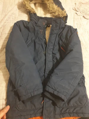 Mörkblå parkajacka från H&M - Parkajacka i mörkblått från H&M med mysigt fuskpälsfoder i luvan och orange detaljer vid dragkedjorna. Jackan har stora fickor framtill, ribbade muddar och dragkedja med knappslå. Perfekt för kalla dagar och har en cool streetstil.