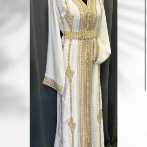 Vit kaftan med guldiga broderier - Elegant vit långklänning med detaljerade guldiga broderier längs hela klänningen och på ärmsluten. Klänningen har ett vackert bälte i midjan och vida långärmar. Materialet känns lyxigt och har en mjuk, glansig yta som ger en exklusiv känsla. Medkommer även matchande hijab. Aldrig använd. Passat xs/s/m