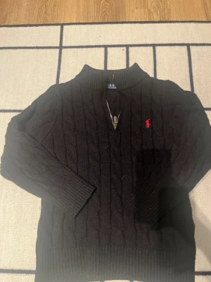 Svart stickad tröja Polo Ralph Lauren - Svart kabelstickad tröja från Polo Ralph Lauren med halv dragkedja och röd broderad logga på bröstet. Tröjan har ribbade muddar och hög krage, perfekt för kyliga dagar. Klassisk och stilren design som funkar till många outfits.