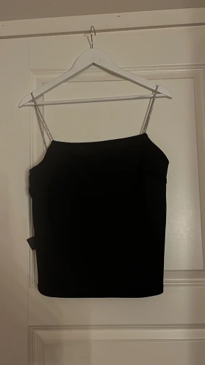 Svart linnetopp med glittriga band - Säljer en stilren svart linnetopp med raka linjer och smala glittriga axelband. Toppen har en enkel och elegant design som passar perfekt till en snygg outfit. Materialet känns mjukt och följsamt mot huden.