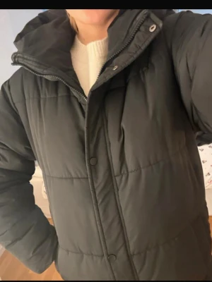 Svart lång pufferjacka från Nelly  - Säljer en svart, lång pufferjacka med luva. Jackan har dragkedja och tryckknappar framtill, samt långa ärmar. Perfekt för kalla dagar och har en loose passform som gör den extra bekväm. Skulle säga att den hade passat en M också. Knappast använd. Kan gå ner i pris 