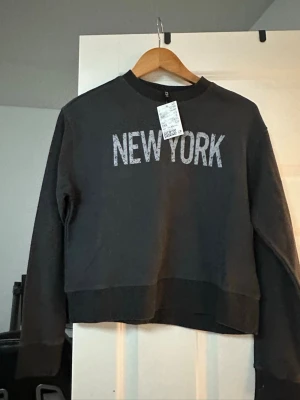 Svart croppad sweatshirt med tryck - Cool svart croppad sweatshirt från H&M med stort vitt 'NEW YORK'-tryck på bröstet. Tröjan har rund hals, långa ärmar och breda muddar vid ärmslut och nederkant. Perfekt för dig som gillar streetstyle och vill ha en avslappnad look.