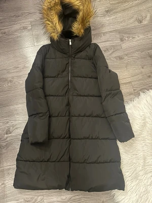 Svart pufferjacka med fuskpäls. Nyskick  - Säljer en svart pufferjacka med huva och snygg fuskpäls.  Skulle jag säga att den är i nyskick då jag endast använt den 2 ggr. Hittar inga fel på den. Perfekt för kalla dagar då den är jättevarm. Avtagbart bälte i midjan.  Nypris 999kr 