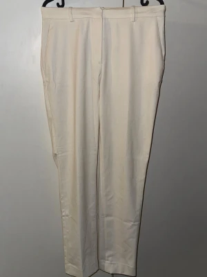 Beiga kostymbyxor från H&M, strl 36 - Snygga beiga kostymbyxor från H&M i storlek 36. Byxorna har raka ben, midjehällor och fickor både fram och bak. Materialet är mjukt och har en lätt struktur, perfekt för en clean och stilren look.