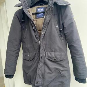 Svart parkajacka från Svea,s-m - Säljer en svart parkajacka från Svea i bra skick. Jackan har en varm vaddering och en praktisk huva. Den har dragkedja och knappar framtill samt två stora fickor med knappar. Perfekt för kalla vinterdagar! 🇺🇸 storlek passar 36-38(40)
