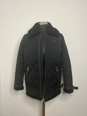 Svart quiltad jacka Peak Performance XS - Quiltad svart jacka från Peak Performance med teddykrage och detaljer i teddy vid ärmslut och nederkant. Jackan har två snedställda fickor med dragkedja, ribbade muddar och snygga metallknappar. Perfekt för kalla dagar med sin stilrena look.