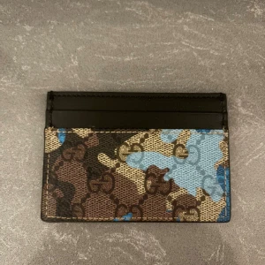 Gucci plånbok med camouflagemönster - Säljer en snygg plånbok från Gucci med camouflagemönster i brunt, beige och blått. Plånboken har flera fack för kort och är tillverkad i skinn med klassiska GG-loggor över hela framsidan. Perfekt för dig som vill ha något stilrent och unikt i fickan.