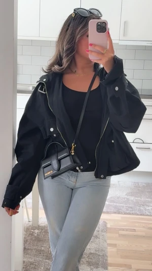 Svart bomberjacka från Jard de Pan - Svart bomberjacka med coola detaljer som silverfärgad dragkedja, stora fickor med knappar och spänne vid kragen. Jackan har en loose passform och är perfekt att slänga över en outfit för en trendig look. Materialet känns som polyester och jackan är långärmad.