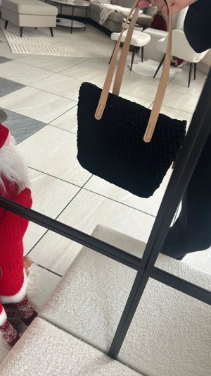 Svart stickad handväska med skinnhandtag - Säljer en svart stickad handväska med snygga ljusa skinnhandtag. Väskan har en rektangulär form och är rymlig, perfekt för att bära med sig allt man behöver. Den stickade ytan ger en cool och unik look som sticker ut.