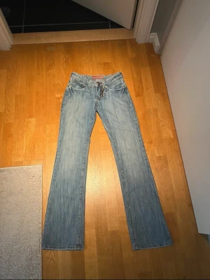 Blå bootcut jeans från D*KLIC - Säljer ett par blå bootcut jeans från D*KLIC med låg midja och snygga dragkedjedetaljer på bakfickorna. De är Lågmidjade och as snygga men tyvärr för små, knappt använda. Midje mått 30cm rakt över och innerbenslängd 78cm
