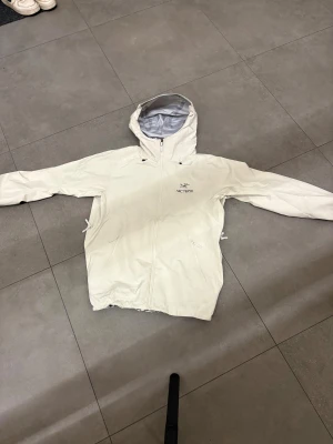 Vit Arc'teryx vindjacka med huva - Säljer en stilren vit vindjacka från Arc'teryx med huva och hel dragkedja framtill. Jackan har två sidofickor med dragkedja och justerbar nederkant. Materialet är lätt och syntetiskt, perfekt för blåsiga dagar. Jackan är enkel att packa ihop och ta med sig. Är perfekt för skidor då den har större luva!