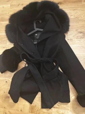 Svart kappa med pälsdetaljer från Amisu - Säljer en svart kappa från Amisu med fluffig päls på huva och ärmslut. Jackan har knytband i midjan och snygga knappar framtill. Perfekt för dig som vill ha en stilren och varm look under höst och vinter.(päls ifrån garoff som jag sytt på därför högre värde) 