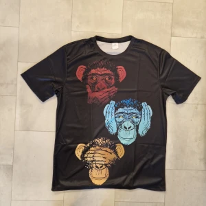 Svart t-shirt med aptryck - Cool svart t-shirt i polyester med tryck av tre apor i rött, blått och gult på både fram- och baksidan. Polyester. T-shirten har rund hals och korta ärmar, perfekt för dig som gillar unika och färgstarka motiv.