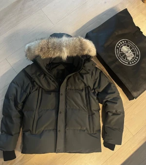 Canada Goose jacka grå - Canada Goose pufferjacka i svart med stor fluffig päls på huvan. Jackan har quiltad design, dragkedja och knappar framtill samt ribbade muddar vid ärmslut. Perfekt för kalla vinterdagar och har en riktigt schysst look. Storlek: L