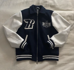 Collegejacka från Replay - Snygg jacka från Replay i mörkblått med vita ärmar och broderade detaljer. Jackan har dragkedja, ribbade muddar med ränder och quiltat foder. Endast använd en gång så jättefint skick! Storlek M från herravdelningen, sitter snyggt oversized på en XS eller S. 