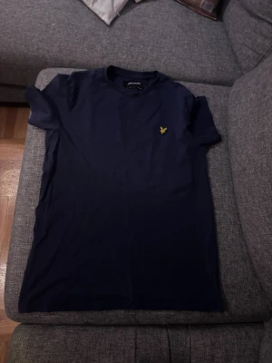 Marinblå t-shirt från Lyle & Scott - Snygg marinblå t-shirt från Lyle & Scott med klassisk rund hals och korta ärmar. På bröstet sitter den ikoniska gula fågeln broderad. T-shirten är gjord i mjuk bomull och har en clean, stilren look som funkar till jeans eller shorts.