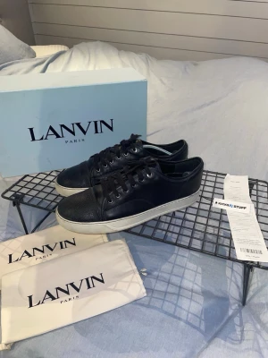Svarta Lanvin sneakers i skinn - Snygga svarta sneakers från Lanvin i äkta skinn med vit platt sula och rund tå. Skorna har snörning och en texturerad tådel som ger en exklusiv känsla. Kommer med originalbox, dustbags och även kvitto. Perfekta för dig som gillar stilrena och lyxiga sneakers.