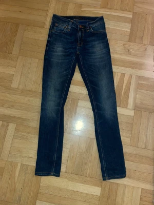 Blå skinny/slim jeans från Nudie Jeans - Snygga blå jeans från Nudie Jeans med klassisk femficksdesign och kontrastsömmar. Modellen är skinny/slim med smal passform och har en mörk tvätt med lätt slitning. Jeansens material är bomull och de har en normal midja. Perfekt för dig som gillar stilrena och tidlösa jeans.