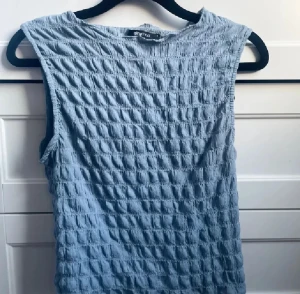 Blå ärmlös topp från Gina Tricot - Säljer en ljusblå topp från Gina Tricot i storlek S. Toppen är ärmlös och har en cool ribbad struktur med rutigt mönster i tyget. Materialet känns mjukt och luftigt, perfekt för varmare dagar. Enkel och stilren design som är lätt att matcha.