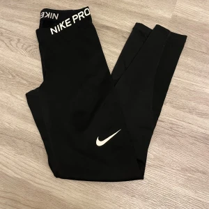 Nike Pro svarta leggings - Svarta Nike Pro leggings med vit logga på benet och bred resår med Nike Pro-tryck i midjan. Snygg, tight passform som sitter skönt och är perfekt för träning. Materialet är stretchigt syntet som andas och ger bra rörelsefrihet.