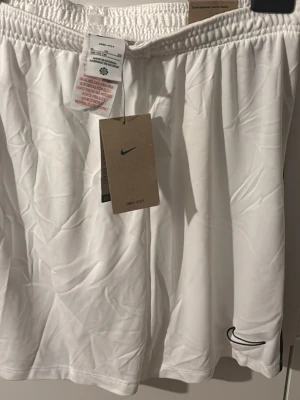 Vita Nike Dri-Fit shorts XL - Säljer ett par vita Nike Dri-Fit shorts i storlek XL. De har elastisk midja och svart Nike-logga broderad på benet. Materialet är lätt polyester och passar perfekt för fotboll eller träning. Snygg och enkel design med svart detalj längs sidan. Dom har också vart i mitt garderob utan användning.