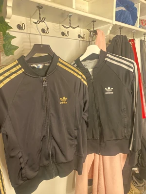 Svart Adidas ziptröja med guld och silver - Två svarta Adidas ziptröjor med klassiska ränder på ärmarna, en med guld och en med silver. Båda har Adidas logga på bröstet och dragkedja framtill. Tröjorna har ribbade muddar och är tillverkade i polyester, perfekt för en sportig och chill stil.