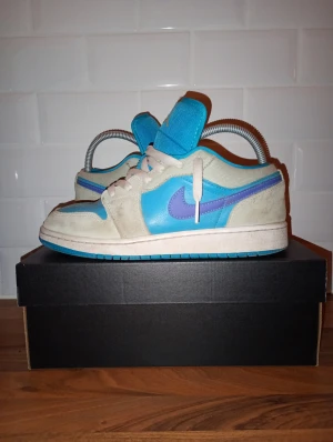 Jordan 1 Low - Nike Jordan 1 Low sneakers i fräsch blå och vit färg med lila swoosh. Skorna har snörning, platt sula och rund tå. Materialet är en mix av skinn och syntet, med en cool kontrast mellan de olika färgpanelerna. Perfekt för dig som gillar streetstyle och vill sticka ut. Vid köp av varan gratis marmor armband!