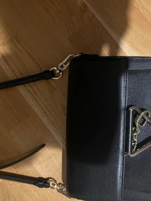 Svart axelväska från Karl Lagerfeld - Snygg svart axelväska från Karl Lagerfeld i stilren design. Väskan är i skinn med silverfärgade metalldetaljer och justerbar axelrem. Framsidan har en dekorativ kedja och logga, och baksidan är enkel med diskret logotyp. Perfekt storlek för det viktigaste.