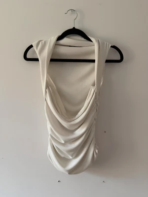 Vit omlott linnetopp med drapering - Säljer en vit linnetopp med breda axelband och snygg draperad front. Toppen har omlott-detalj och är ärmlös, vilket ger en cool och modern look. Materialet är mjukt och har ett lätt glansigt utseende som känns lyxigt. Perfekt att styla med jeans eller kjol.