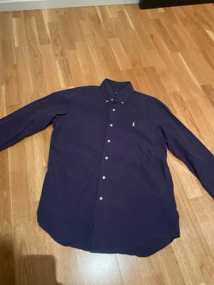 Mörkblå skjorta från Ralph Lauren - Säljer en stilren mörkblå skjorta från Ralph Lauren i storlek M. Skjortan har klassisk krage, långa ärmar och knappar framtill. På bröstet finns den ikoniska broderade loggan i gult. Materialet är mjuk bomull som känns skönt mot huden.