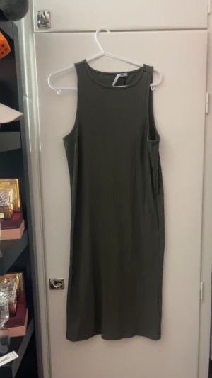Ribbad grön långklänning från 157 - Säljer en stilren långklänning i ribbad grön bomull från 157. Klänningen är ärmlös med rund hals och har en loose passform som ger en avslappnad vibe. Perfekt för dig som gillar enkel och bekväm stil.