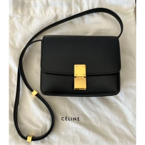 Svart axelväska från Céline i skinn - Stilren svart axelväska från Céline i slätt skinn med guldigt lås och detaljer. Väskan har en klassisk, fyrkantig form och justerbar axelrem. Insidan har flera fack för smart organisering. Perfekt för dig som gillar minimalistisk och lyxig design.