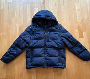 Mörkblå vinterjacka från Polo Ralph Lauren - Säljer en mörkblå pufferjacka från Polo Ralph Lauren med huva och dragkedja framtill. Jackan är använd 1 säsong och har tre fickor med dragkedja och ett rött Polo-logo broderat på bröstet. Perfekt till en julklapp eller en u vinterjacka 