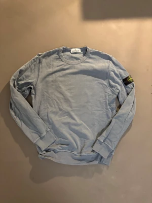 Ljusblå långärmad tröja Stone Island - Snygg ljusblå långärmad tröja från Stone Island med klassisk rund hals och ikonisk patch på vänster ärm. Tillverkad i mjuk bomull som är skön mot huden. Perfekt för dig som gillar stilrena och avslappnade plagg med streetkänsla.