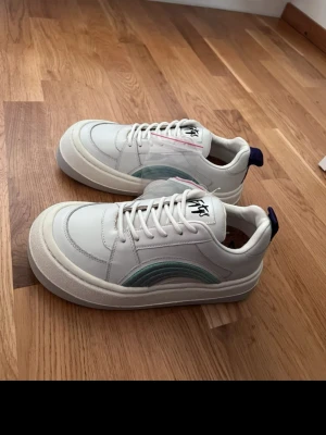 Vita Eytys sneakers med blå detalj - Säljer ett par vita sneakers från Eytys med coola blå och gröna vågdetaljer på sidan. Skorna har tjock platt sula, rund tå och snörning. Märkets logga syns på plösen och det finns en mörkblå hälflik. Materialet är skinn och syntet.