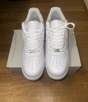 Nike Air Force 1 helvita sneakers - Klassiska Nike Air Force 1 sneakers i helvitt skinn med perforerad tå, rund tå och platt sula. Skorna har snörning och diskret Nike-logga på sidan. Perfekta för en clean och stilren look.