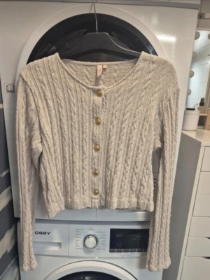 Beige stickad kofta med guldknappar - Säljer en beige stickad kofta från Nelly med snygga guldknappar framtill. Koftan har ett ribbat och kabelstickat mönster, lång ärm och croppad passform. Perfekt att slänga över en t-shirt eller linne för en chill look.