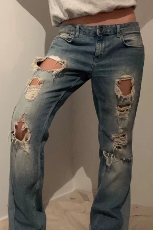 Blå ripped jeans med loose fit - Säljer ett par ljusblå jeans med rejält slitna och trasiga detaljer på båda benen. Modellen har loose passform och raka ben. Midjemått rakt över: 42 cm Innerbenslängd: 82cm