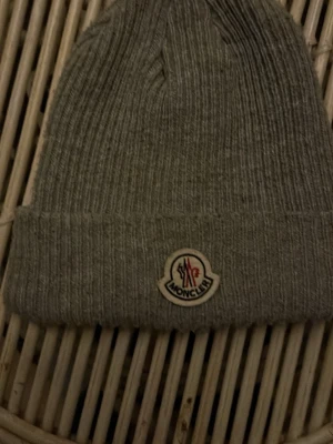 Grå ribbstickad Moncler mössa - Säljer en snygg grå mössa från Moncler med ribbstickad design och uppvikt kant. Mössan har Moncler-loggan framtill och är tillverkad i mjukt material som håller dig varm. Perfekt för dig som vill ha en stilren accessoar med exklusiv känsla.