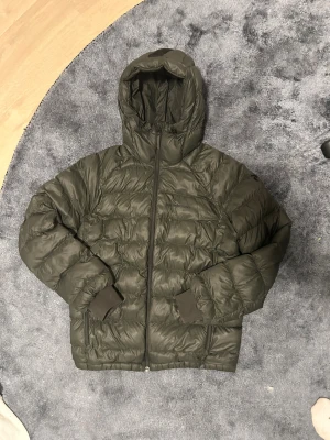 Mörkgrön  dunjacka från Peak Performance - Mörkgrön dunjacka från Peak Performance med huva och dragkedja framtill. Jackan har quiltad design, ribbade muddar vid ärmslut och en snygg rem på huvan. Perfekt för kalla dagar och har en clean, sportig look.