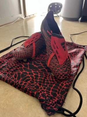 Adidas Predator Year of the Snake fotbollsskor - Säljer ett par Adidas Predator fotbollsskor med unikt 'Year of the Snake'-tema. Skorna har svart och röd ormmönstrad design, guldiga detaljer på sulan och röda snören. Tillverkade i syntetmaterial med platt sula och rund tå. Perfekt för dig som vill sticka ut på planen. Defekt på höger skon men man märker inte av ngt när man spelar om man inte tänker på de.