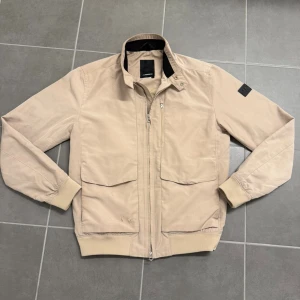 Beige vindjacka från J.Lindeberg M - Snygg beige vindjacka från J.Lindeberg med klassisk ståkrage och svarta detaljer på insidan av kragen. Jackan har två stora fickor framtill, ribbade muddar och dragkedja. Perfekt för dig som gillar stilren design och vill ha något lätt att slänga på sig.