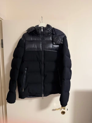 Svart pufferjacka Polo Ralph Lauren - Mörkblå pufferjacka från Polo Ralph Lauren med glansiga detaljer upptill och på huvan. Jackan har två dragkedjefickor fram, hel dragkedja och en stor, vadderad huva. Materialmix med polyester och syntet, perfekt för kalla dagar.