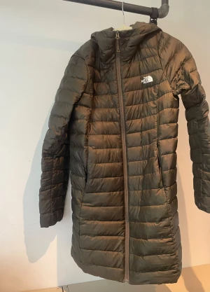 Lång dunjacka från The North Face - Snygg lång dunjacka från The North Face i mörkgrön färg. Jackan har huva, dragkedja hela vägen och quiltad design. Perfekt för kyliga dagar och outdoor-aktiviteter. Logga på bröstet och lätt vadderad för extra värme.