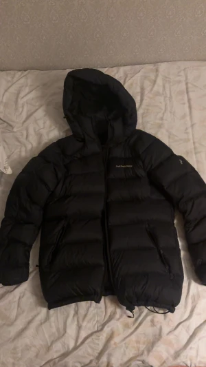 Svart dunjacka från Peak Performance - Säljer en svart dunjacka från Peak Performance med huva och dragkedja. Jackan har en puffig look, två sidofickor med dragkedja och diskret logga på bröstet. Perfekt för kalla dagar och riktigt snygg till streetwear-stilen. Storleken finns i både M och S