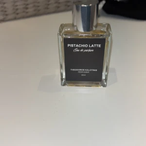 Pistachio Latte Eau de Parfum 50ml - Unik parfym med doft av pistage och latte från Theodoros Kalotinis. Flaskan är rektangulär i genomskinligt glas med en stilren svart etikett och silverfärgad kork. Innehåller 50 ml Eau de Parfum – perfekt för dig som gillar gourmanddofter.