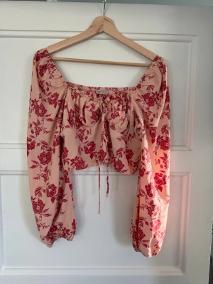 Blommig croppad blus från Glamorous - Säljer en ljusrosa croppad blus från Glamorous med röda blommor, puffiga långärmar och snörning framtill. Blusen har en fyrkantig urringning och smockad rygg för extra komfort. Perfekt för dig som gillar romantiska och trendiga detaljer.