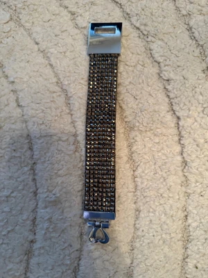 Glittrigt brett armband med hjärtdetalj - Snyggt brett armband med glittrande stenar i silverton och rutigt mönster. Spännet har en söt hjärtdetalj med liten sten. Baksidan är svart och slät, medan framsidan är täckt av små fyrkantiga stenar som ger extra bling. Perfekt för dig som gillar att sticka ut.