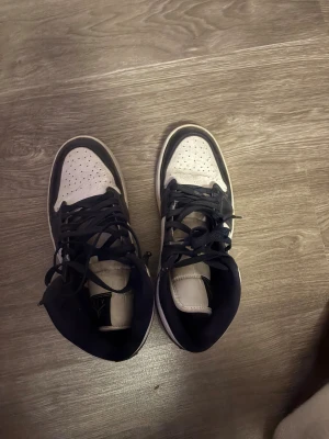 Nike Air Jordan 1 svart/vit sneakers - Säljer ett par Nike Air Jordan 1 sneakers i svart och vitt med klassisk high-top siluett. Skorna har snörning, perforerad tå och ikoniska Air Jordan-loggan på sidan. Materialet är skinn och de har en platt sula med rund tå. Perfekt för dig som gillar streetstyle och vill sticka ut.