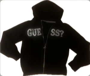 Svart hoodie från Guess med paljetter - Snygg svart hoodie från GUESS – dammodell  Superfin svart zip-hoodie med glittrande silverpaljetter som bildar texten “GUE SS?” på bröstet. Bekväm passform, mjukt material och huva med dragkedja som går hela vägen ner. Klassisk och samtidigt iögonfallande – perfekt både till vardag och mer uppklädda casual-outfits.  Skick: Mycket bra begagnat skick, inga fläckar eller hål. Detaljer: 	•	Hel dragkedja framtill 	•	Kängurufickor 	•	Paljettbrodyr 	•	Mjuk och varm kvalitet  Perfekt för dig som gillar 